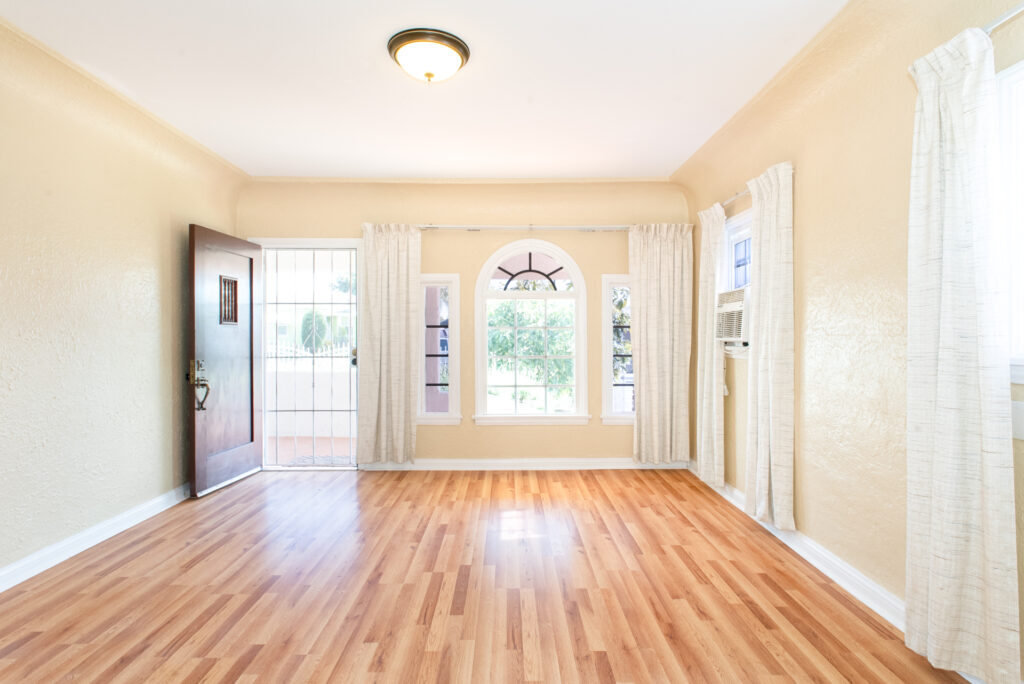 Bright living room with wood floors, arched window and open front door, photographed wide for real estate listings.
